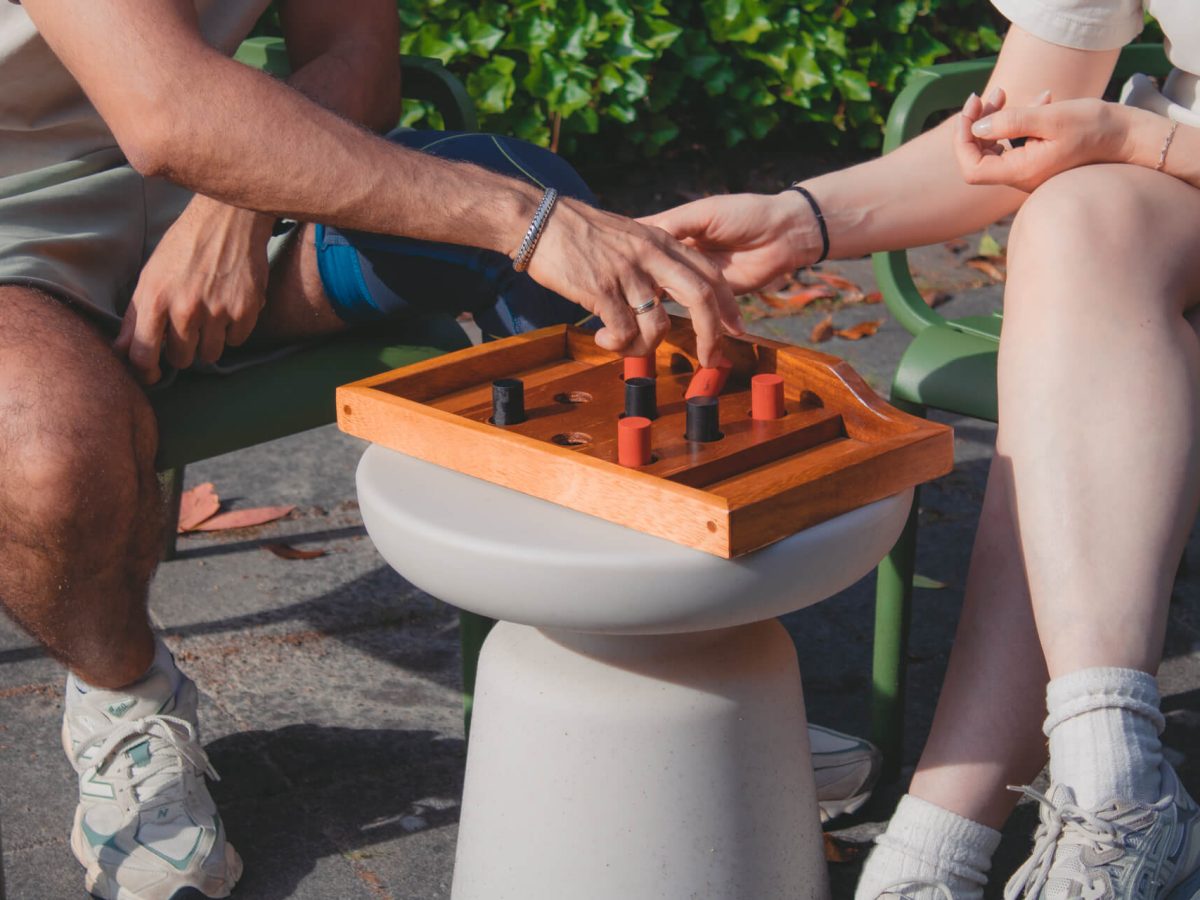 Twee personen zitten op stoelen en spelen het klassieke spel boter, kaas en eieren op een klein bijzettafeltje. Alleen hun benen zijn zichtbaar.